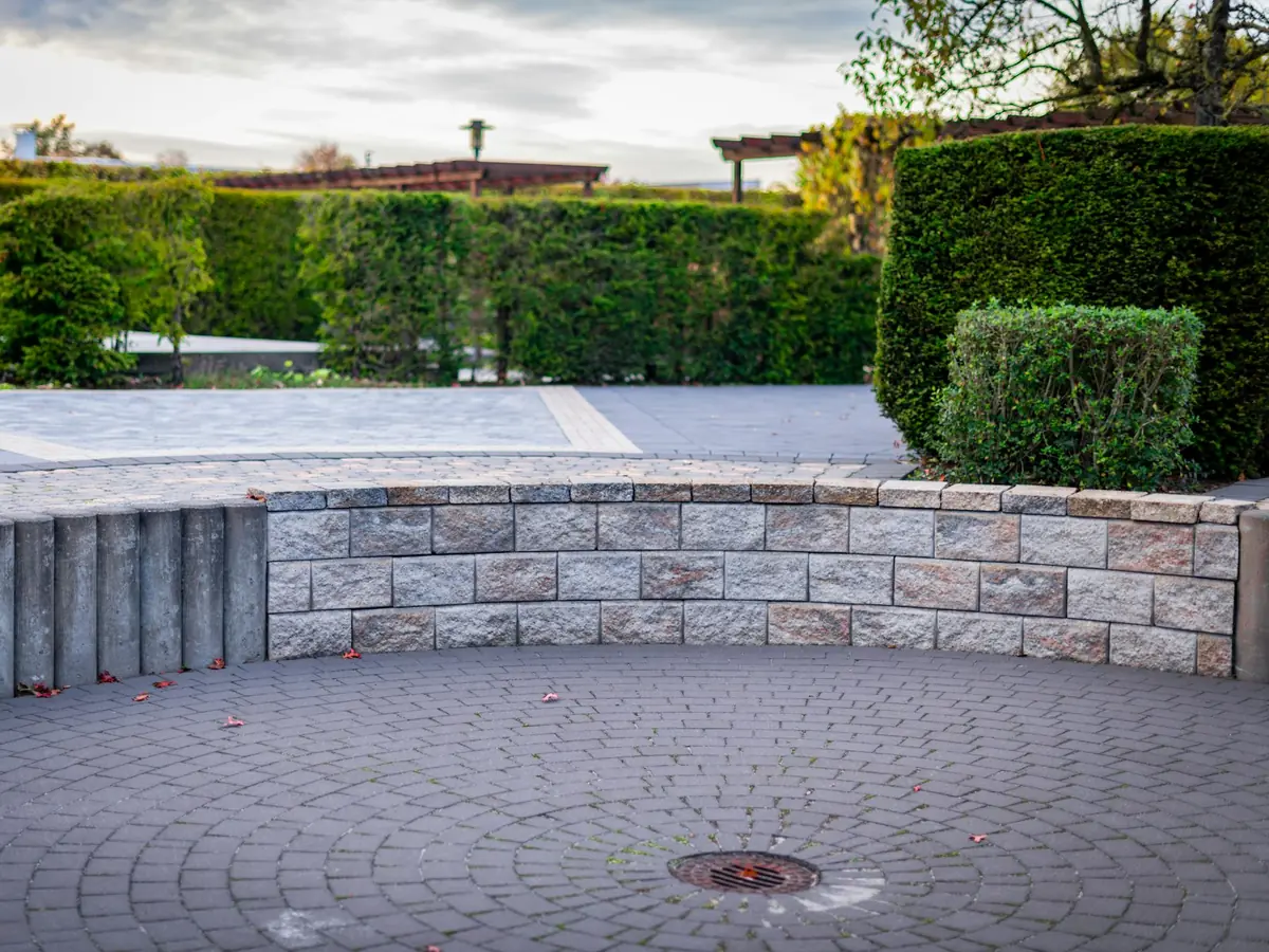Terrasse pavée en arc de cercle bordée par un muret en pierre et une haie taillée