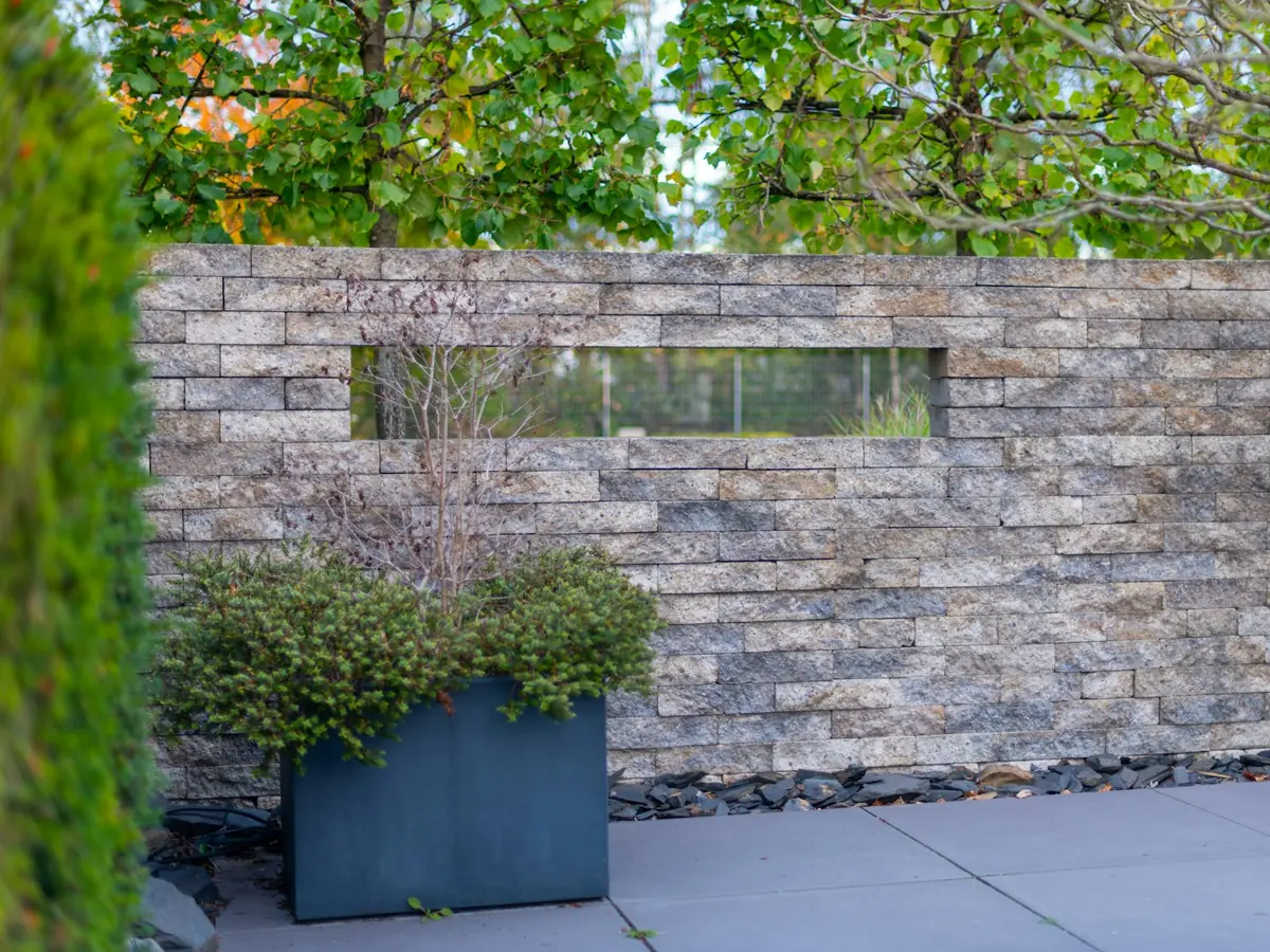Mur en pierre avec un bac en métal planté devant une allée pavée